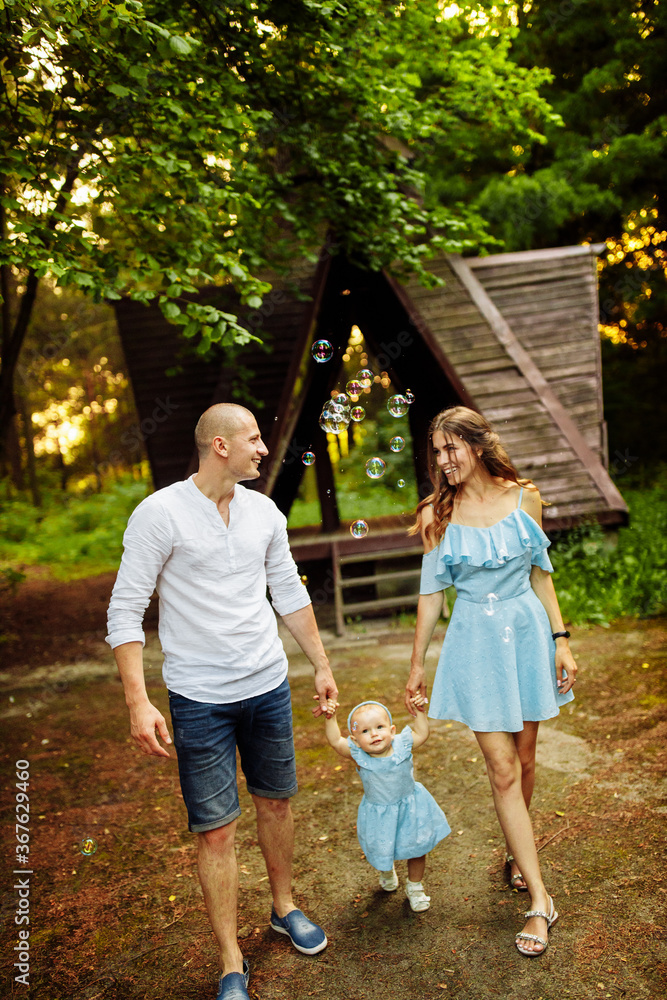 Fototapeta premium Happy young family walking at the park, cute toddler looking at bubbles, lovely daughter hold mother and father hands, mom and dad looking at each other, smiling, parenting concept