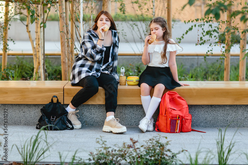 Two schoolgirls, a child and a teenager, have their lunch on a bench in the schoolyard.