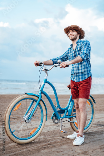 Smiling boy with his bike in a straw hat, standing on the beach with the sea on the backgroung