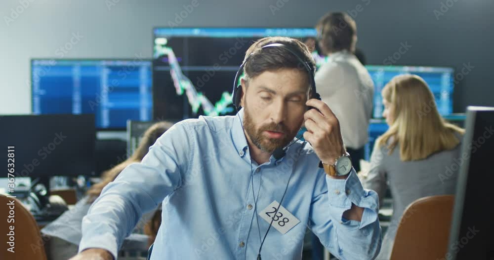 Caucasian handsome male broker in headset working at computer in trading office and answering phone calls on many telephones. Trader buying and selling while talking with clients and working hard. Caucasian handsome male broker in headset working at computer in trading office and answering phone calls on many telephones. Trader buying and selling while talking with clients and working hard.