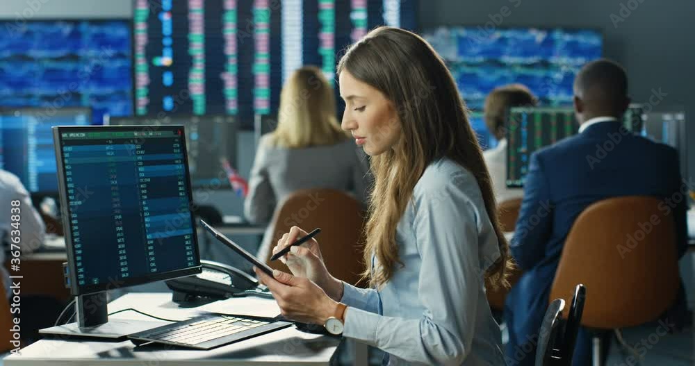 Caucasian woman broker sitting in front of computer monitor with charts and graphics and noting in tablet device. Female trader buying and selling stocks. Putting bets and rates. Analyzing market. Caucasian woman broker sitting in front of computer monitor with charts and graphics and noting in tablet device. Female trader buying and selling stocks. Putting bets and rates. Analyzing market.