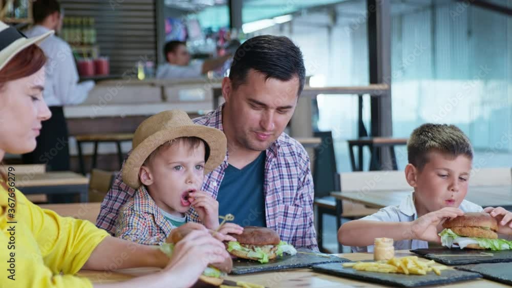 happy cheerful man and woman with male children enjoy eating tasty juicy burgers with fries sitting at table in cafe happy cheerful man and woman with male children enjoy eating tasty juicy burgers with fries sitting at table in cafe