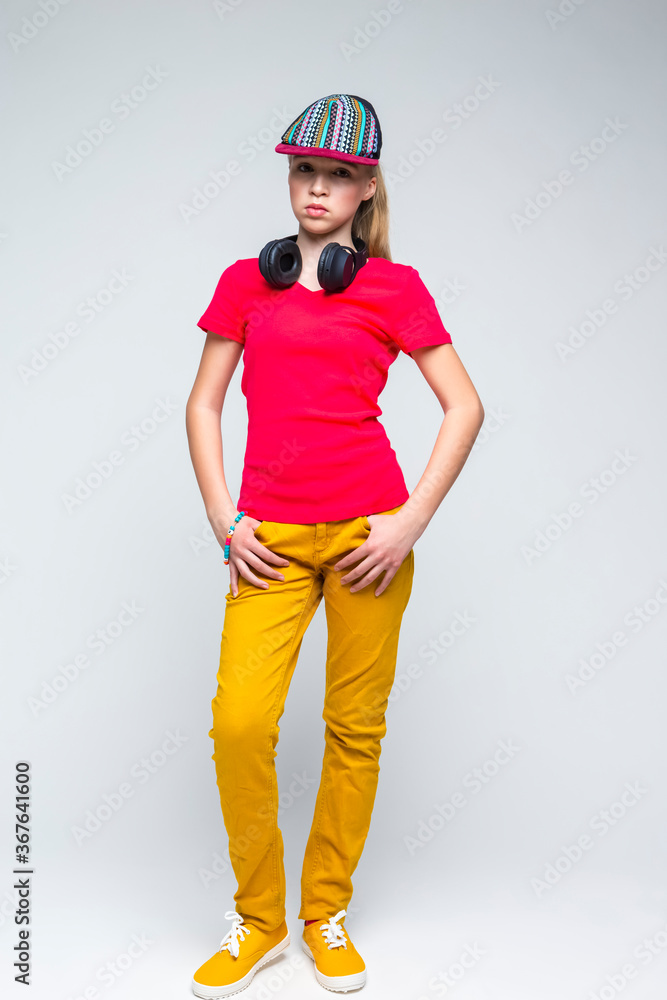 Youth Lifestyle. Full Length Portrait of Tranquil Caucasian Teenage Girl with Wireless Headphones. Posing in Colorful Cap Over Gray Background. Youth Lifestyle. Full Length Portrait of Tranquil Caucasian Teenage Girl with Wireless Headphones. Posing in Colorful Cap Over Gray Background.