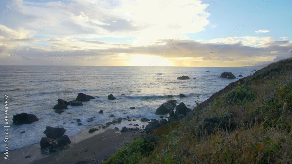 Shot of sun setting over rocky coast bay. Cloudy sky. Grassy hill in foreground. Shot of sun setting over rocky coast bay. Cloudy sky. Grassy hill in foreground.