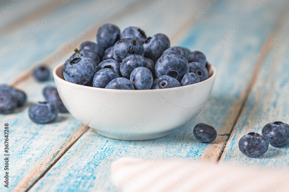 Close up shot of a white bowl of blueberries on light blue, worn, rustic wooden table with some blueberries scattered nearby Close up shot of a white bowl of blueberries on light blue, worn, rustic wooden table with some blueberries scattered nearby