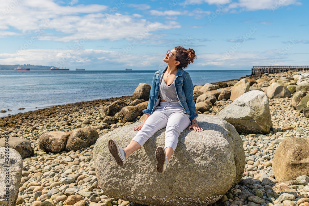 Happy young woman sitting on a big rock at the beach swinging her legs