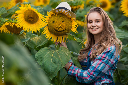 Gorgeous, young, adorable, energetic, female farmer standing near a sunflower with a smiley face, in the middle of a golden sunflower field.
