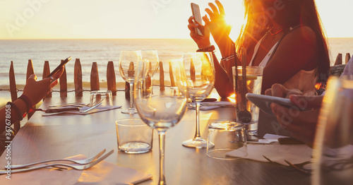 Selective focus on hands holding smartphones while sitting on  restaurant’s terrace during stunning sunset, vintage style. Summer vacation concept