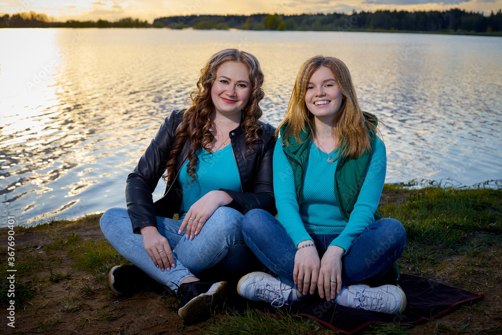 Two girls friends, sisters, cousins socializing and having fun on the shore or beach of a lake or river with calm water and sunset in the background on the horizon. Two girls friends, sisters, cousins socializing and having fun on the shore or beach of a lake or river with calm water and sunset in the background on the horizon.