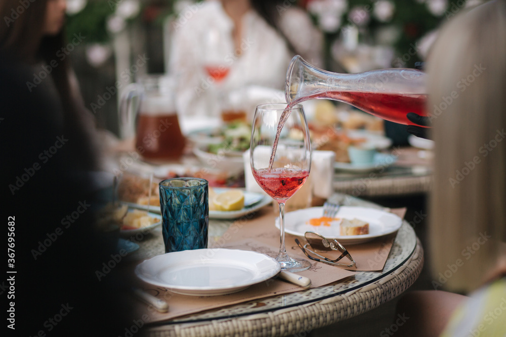 Waiteress pouring rose wine in glass. Background of people eating dinner at summer terrace in cafe. Food concept outdoors. Different kind of food and drinks on the table