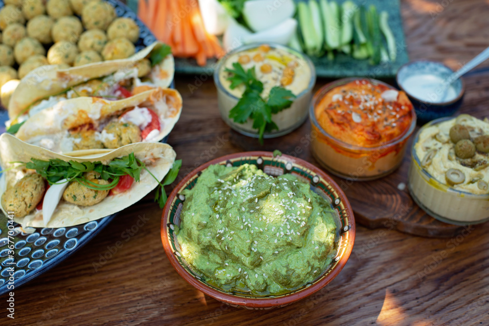 Vegetarian picnic on a wooden oak table, hummus, falafel, pita, vegetables, spinach, basil and sunflower seed pesto sauce. Healthy Mediterranean food. Vegetarian picnic on a wooden oak table, hummus, falafel, pita, vegetables, spinach, basil and sunflower seed pesto sauce. Healthy Mediterranean food.