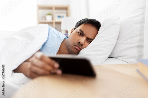 people, bedtime and rest concept - sleepy indian man with smartphone lying in bed at home
