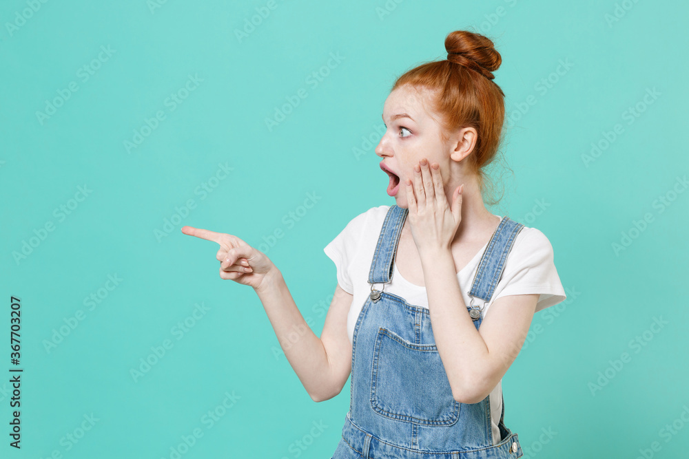 Fototapeta premium Shocked young readhead girl in casual denim clothes posing isolated on blue turquoise background studio. People lifestyle concept. Mock up copy space. Pointing index finger aside, put hand on cheek.