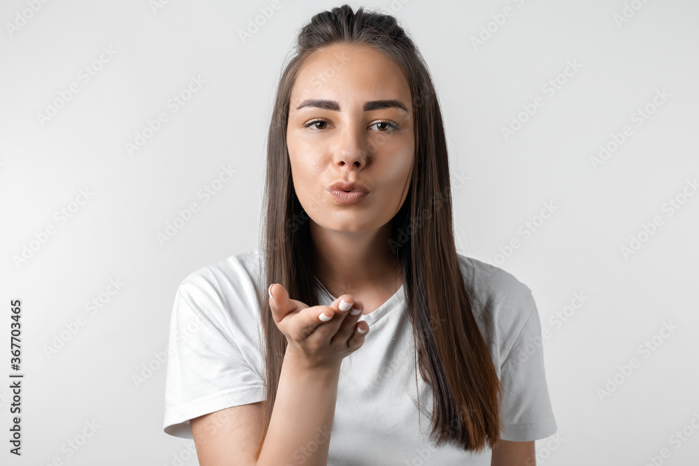 Beautiful European young woman with long chestnut hair makes air kiss, blows over palm, wants to kiss man. Playful female dressed in casual clothes blowing air kiss at camera Beautiful European young woman with long chestnut hair makes air kiss, blows over palm, wants to kiss man. Playful female dressed in casual clothes blowing air kiss at camera