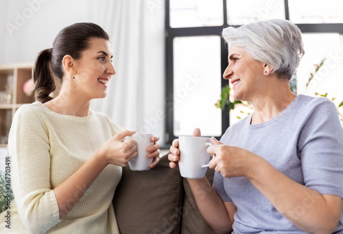 family, generation and people concept - happy smiling senior mother with adult daughter drinking coffee or tea and talking at home