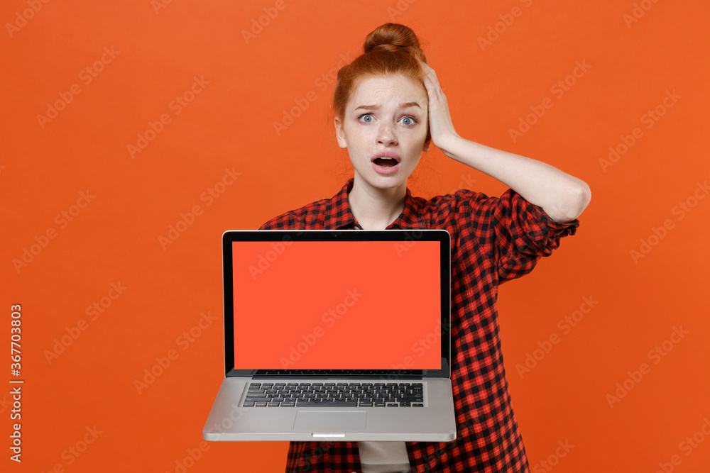 Worried young readhead girl in casual red checkered shirt isolated on orange background. People lifestyle concept. Mock up copy space Hold laptop pc computer with blank empty screen put hand on head. Worried young readhead girl in casual red checkered shirt isolated on orange background. People lifestyle concept. Mock up copy space Hold laptop pc computer with blank empty screen put hand on head.