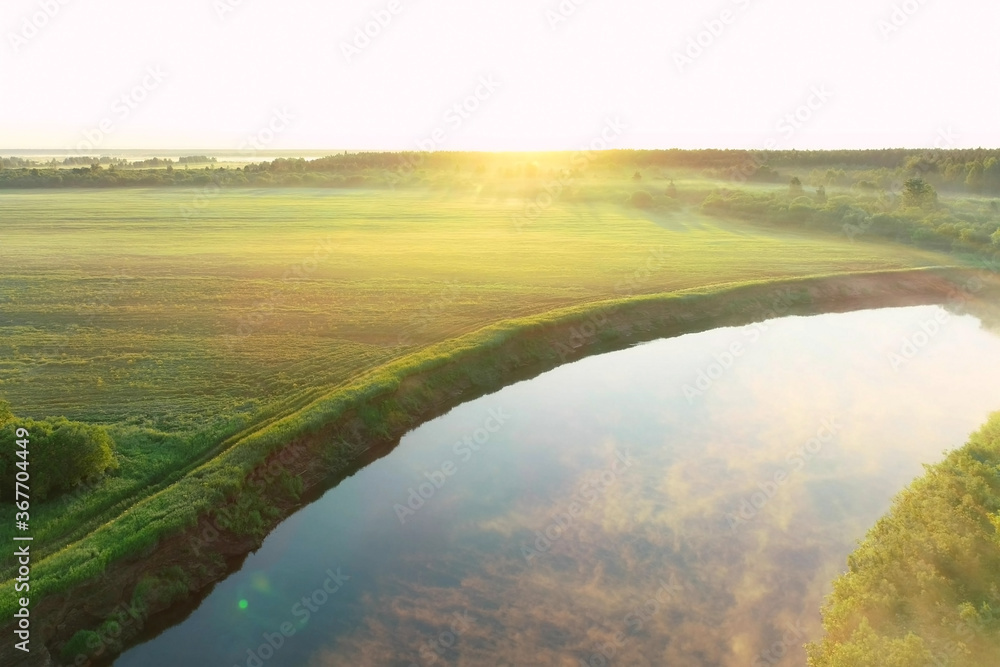 Flying over river with white mist and green forest at summer sunny day, sky reflecting in water. Sunrise on nature in beautiful place with natural landscape. Aerial view of picturesque woodland.