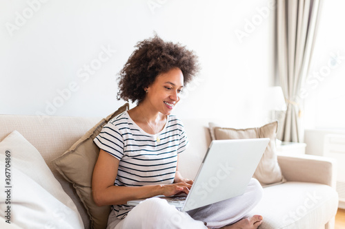 Young beautiful woman with curly hair working on laptop computer while sitting on the sofa at home, heck on oline shops for cyber monday sales. Technology woman concept alternative office freelance photo