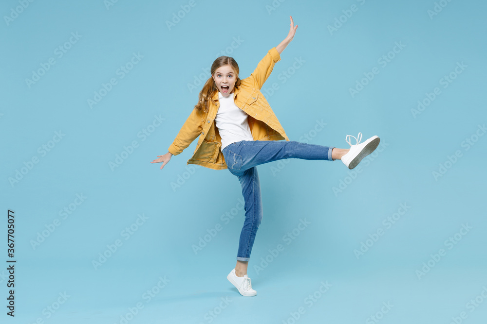 Full length portrait excited little blonde kid girl 12-13 years old in yellow jacket isolated on blue wall background. Childhood lifestyle concept. Mock up copy space. Rising spreading hands and legs.