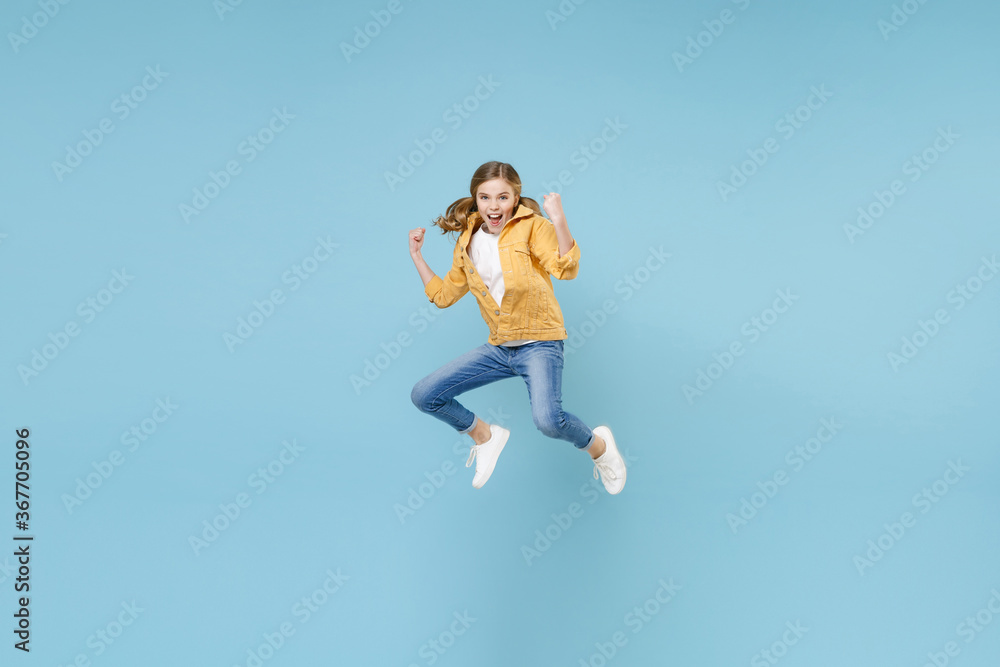 Full length portrait of happy little blonde kid girl 12-13 years old in yellow jacket isolated on blue background studio. Childhood lifestyle concept. Mock up copy space. Jumping doing winner gesture.