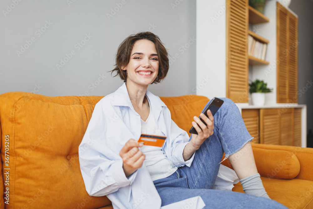 Charming young woman in casual clothes sit on couch spending time in living room at home. Rest relax good mood leisure lifestyle concept. Mock up copy space. Using mobile phone, hold credit bank card. Charming young woman in casual clothes sit on couch spending time in living room at home. Rest relax good mood leisure lifestyle concept. Mock up copy space. Using mobile phone, hold credit bank card.