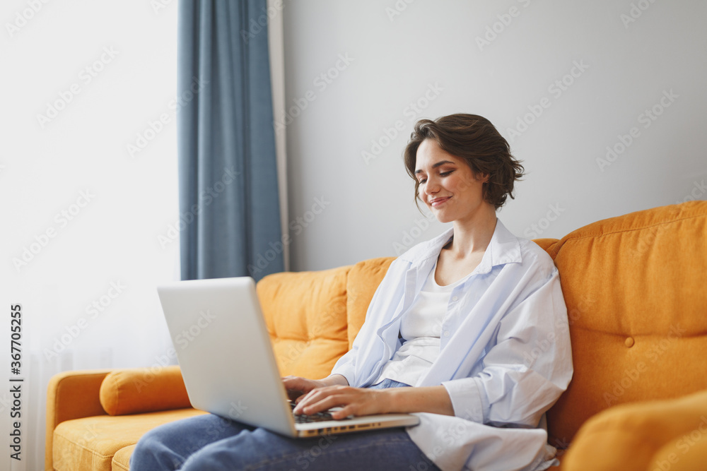 Smiling young woman girl in casual clothes sitting on couch spending time in living room at home. Rest relax good mood leisure lifestyle concept. Mock up copy space. Working on laptop pc computer. Smiling young woman girl in casual clothes sitting on couch spending time in living room at home. Rest relax good mood leisure lifestyle concept. Mock up copy space. Working on laptop pc computer.