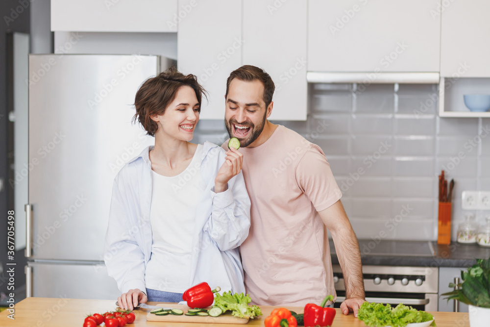 Funny young couple friends guy girl in casual clothes preparing vegetable salad cooking food in kitchen at home. Dieting family healthy lifestyle concept. Mock up copy space. Feeding eating cucumber.