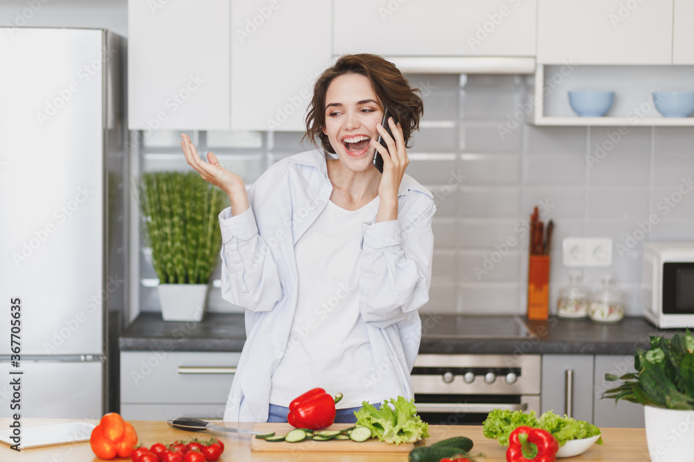 Laughing young housewife woman girl in casual clothes preparing vegetable salad cooking food in light kitchen at home. Dieting healthy lifestyle concept. Mock up copy space. Talking on mobile phone.