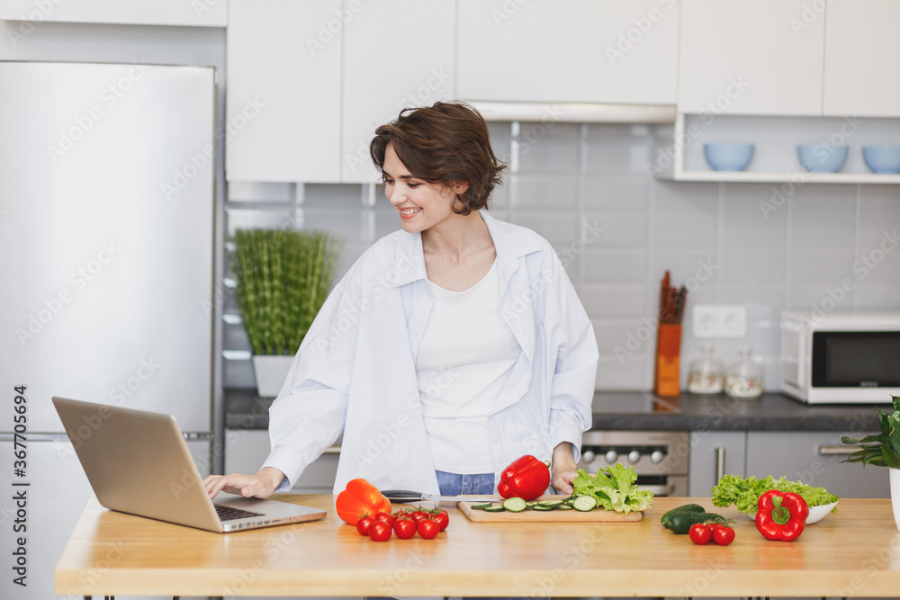 Smiling young housewife woman girl in white casual clothes using laptop computer looking at recipe preparing vegetable salad cooking food in light kitchen at home. Dieting healthy lifestyle concept. Smiling young housewife woman girl in white casual clothes using laptop computer looking at recipe preparing vegetable salad cooking food in light kitchen at home. Dieting healthy lifestyle concept.