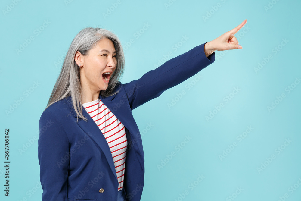 Irritated gray-haired business woman in blue suit isolated on blue background studio. Achievement career wealth business concept. Mock up copy space. Pointing index finger aside up screaming swearing. Irritated gray-haired business woman in blue suit isolated on blue background studio. Achievement career wealth business concept. Mock up copy space. Pointing index finger aside up screaming swearing.