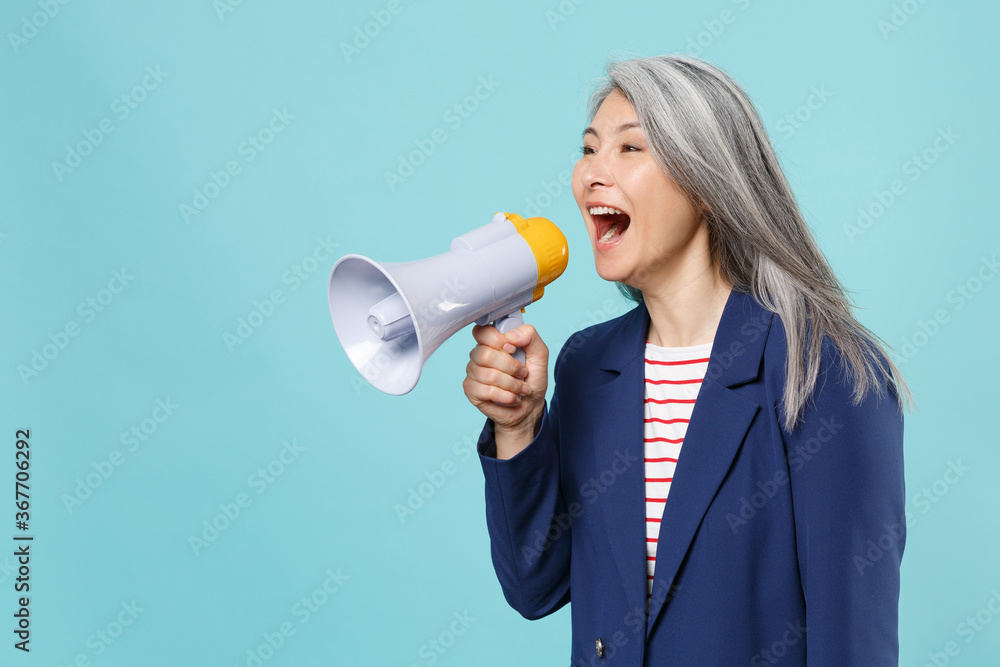 Excited gray-haired business woman in blue suit posing isolated on blue background studio portrait. Achievement career wealth business concept. Mock up copy space. Scream in megaphone, looking aside. Excited gray-haired business woman in blue suit posing isolated on blue background studio portrait. Achievement career wealth business concept. Mock up copy space. Scream in megaphone, looking aside.