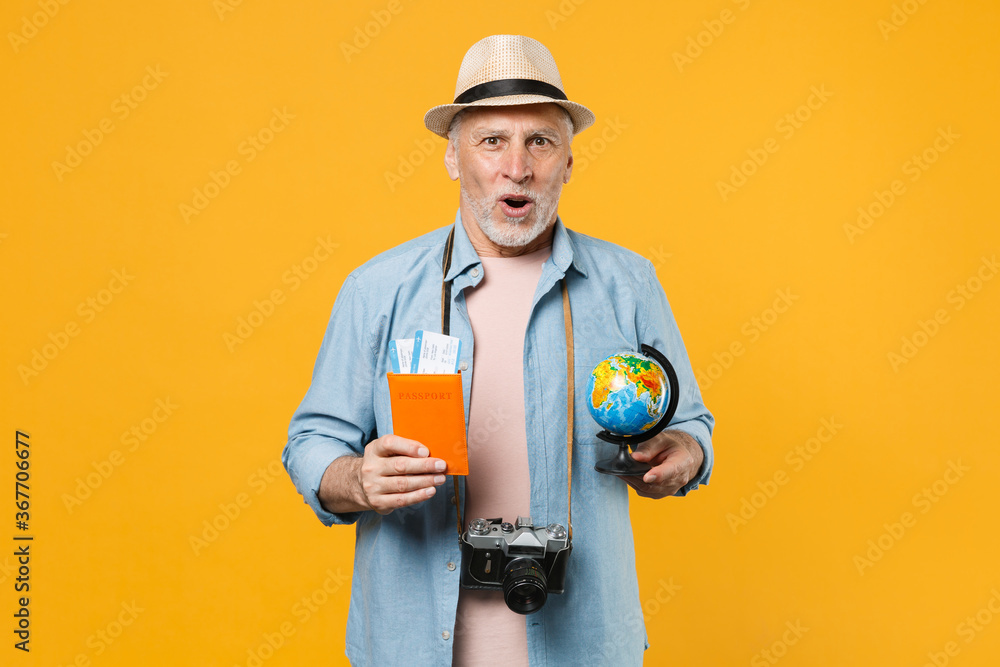 Shocked traveler tourist elderly gray-haired man in hat photo camera isolated on yellow background. Passenger traveling abroad on weekend getaway. Air flight journey. Hold passport ticket world globe.
