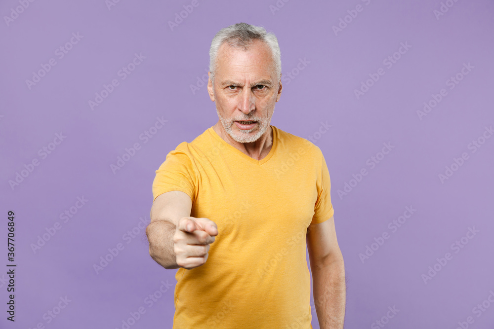 Strict elderly gray-haired mustache bearded man in yellow t-shirt posing isolated on pastel violet wall background. People lifestyle concept. Mock up copy space. Pointing index finger on camera. Strict elderly gray-haired mustache bearded man in yellow t-shirt posing isolated on pastel violet wall background. People lifestyle concept. Mock up copy space. Pointing index finger on camera.