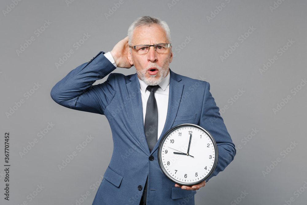 Worried elderly gray-haired bearded business man in classic blue suit shirt tie isolated on grey background studio portrait. Achievement career wealth business concept. Hold clock, put hand on head. Worried elderly gray-haired bearded business man in classic blue suit shirt tie isolated on grey background studio portrait. Achievement career wealth business concept. Hold clock, put hand on head.