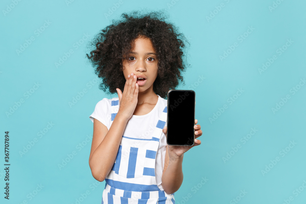 Shocked little african american kid girl 12-13 years old in striped clothes isolated on blue background. Childhood lifestyle concept Hold mobile phone with blank empty screen covering mouth with hand. Shocked little african american kid girl 12-13 years old in striped clothes isolated on blue background. Childhood lifestyle concept Hold mobile phone with blank empty screen covering mouth with hand.