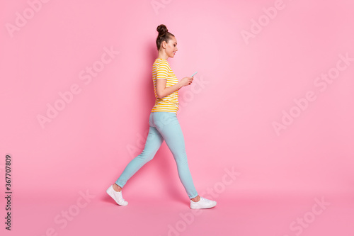 Full length profile photo of attractive lady two funny buns hold telephone hands writing new blog post walk down street wear casual striped t-shirt pants shoes isolated pink color background