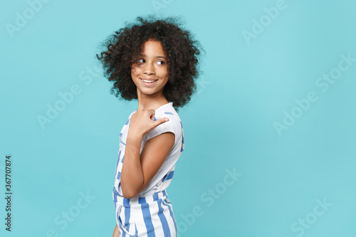 Side view of smiling little african american kid girl 12-13 years old in striped clothes isolated on blue wall background studio. Childhood lifestyle concept. Mock up copy space. Pointing thumb aside.