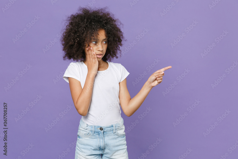 Shocked little african american kid girl 12-13 years old in white t-shirt isolated on violet wall background studio portrait. Childhood lifestyle concept. Pointing index finger up, put hand on cheek. Shocked little african american kid girl 12-13 years old in white t-shirt isolated on violet wall background studio portrait. Childhood lifestyle concept. Pointing index finger up, put hand on cheek.