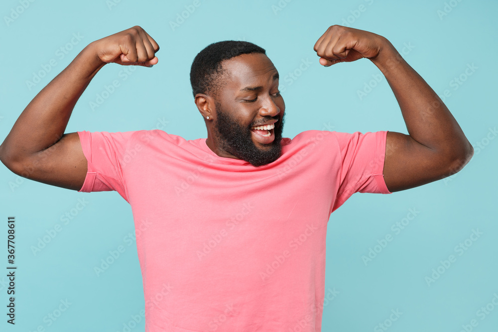 Smiling young african american man guy in casual pink t-shirt posing isolated on pastel blue background studio portrait. People emotions lifestyle concept. Mock up copy space. Showing biceps, muscles. Smiling young african american man guy in casual pink t-shirt posing isolated on pastel blue background studio portrait. People emotions lifestyle concept. Mock up copy space. Showing biceps, muscles.