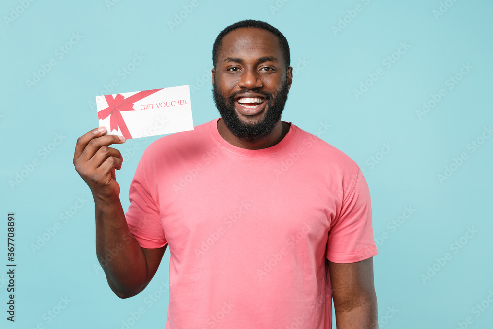 Smiling young african american man guy in casual pink t-shirt isolated on blue wall background studio portrait. People sincere emotions lifestyle concept. Mock up copy space. Hold gift certificate. Smiling young african american man guy in casual pink t-shirt isolated on blue wall background studio portrait. People sincere emotions lifestyle concept. Mock up copy space. Hold gift certificate.