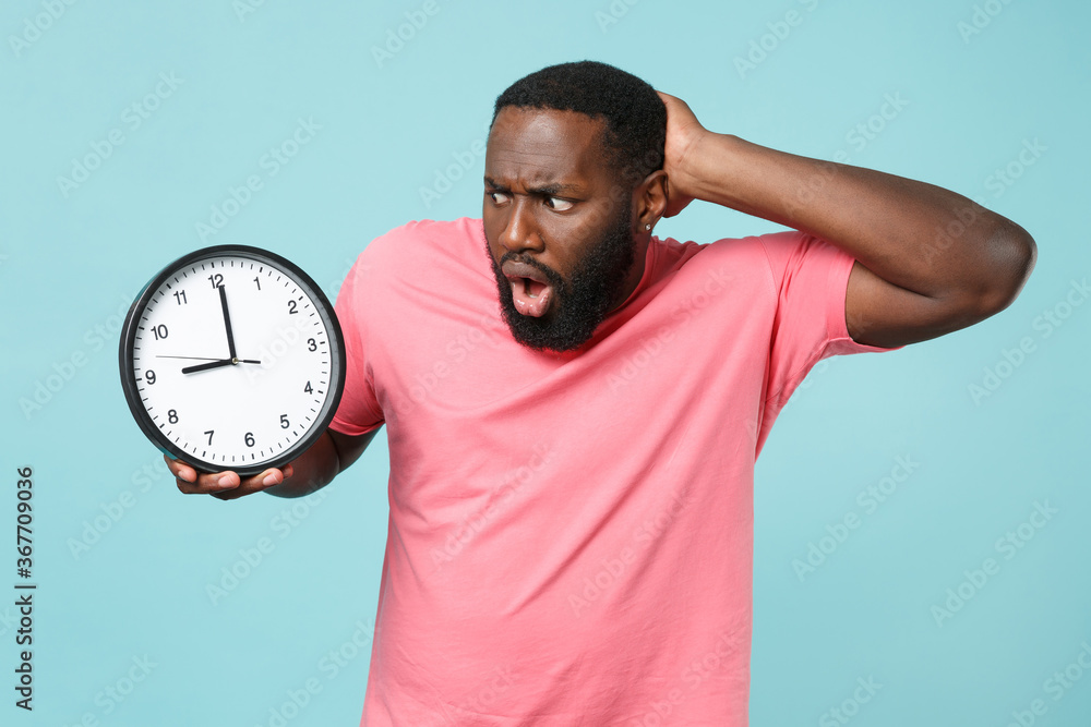 Shocked young african american man guy in casual pink t-shirt isolated on blue background studio portrait. People sincere emotions lifestyle concept. Mock up copy space. Hold clock, put hand on head. Shocked young african american man guy in casual pink t-shirt isolated on blue background studio portrait. People sincere emotions lifestyle concept. Mock up copy space. Hold clock, put hand on head.