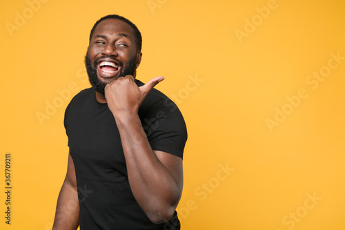 Cheerful african american man football fan in casual black t-shirt isolated on yellow background studio portrait. People sincere emotions lifestyle concept. Mock up copy space. Pointing thumb aside.