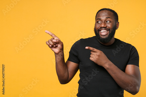 Smiling african american man football fan in casual black t-shirt isolated on yellow background studio portrait. People emotions lifestyle concept. Mock up copy space. Pointing index fingers aside.