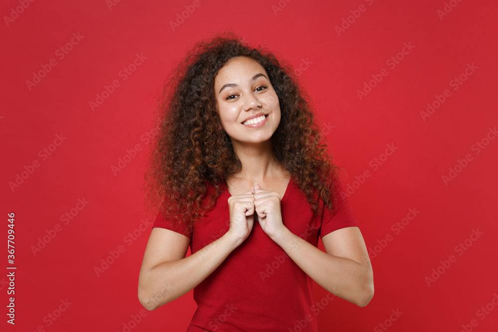 Smiling young african american woman girl in casual t-shirt posing isolated on bright red background studio portrait. People sincere emotions lifestyle concept. Mock up copy space. Clenching fists. Smiling young african american woman girl in casual t-shirt posing isolated on bright red background studio portrait. People sincere emotions lifestyle concept. Mock up copy space. Clenching fists.