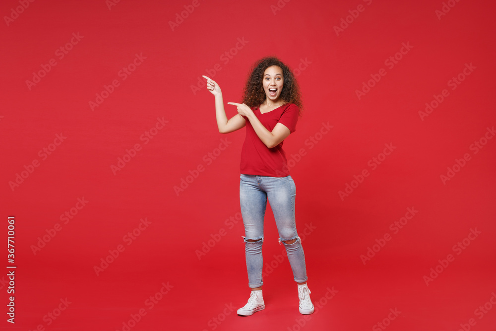 Full length portrait of excited young african american girl in casual t-shirt isolated on red background studio portrait. People lifestyle concept. Mock up copy space. Pointing index fingers aside.