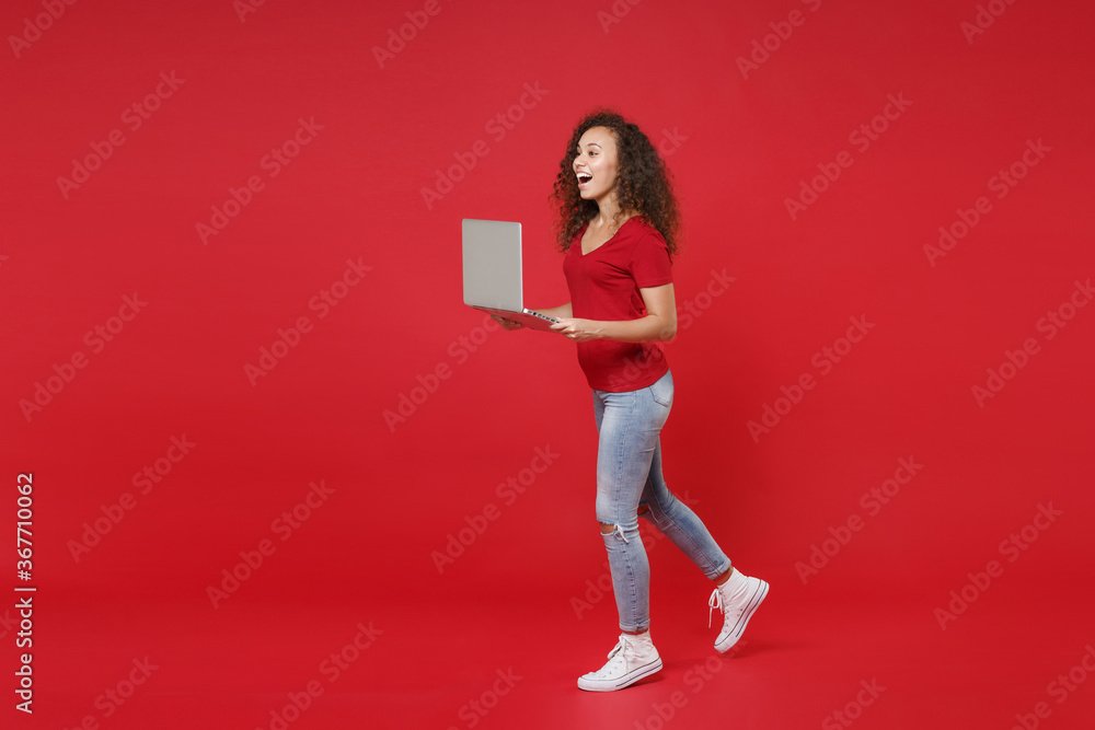 Full length portrait side view excited young african american girl in casual t-shirt isolated on red background studio. People lifestyle concept. Mock up copy space. Running with laptop pc computer. Full length portrait side view excited young african american girl in casual t-shirt isolated on red background studio. People lifestyle concept. Mock up copy space. Running with laptop pc computer.