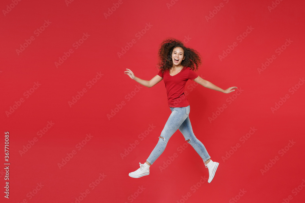 Full length portrait side view of funny young african american girl in casual t-shirt isolated on red background studio. People lifestyle concept. Mock up copy space. Jumping spreading hands and legs.