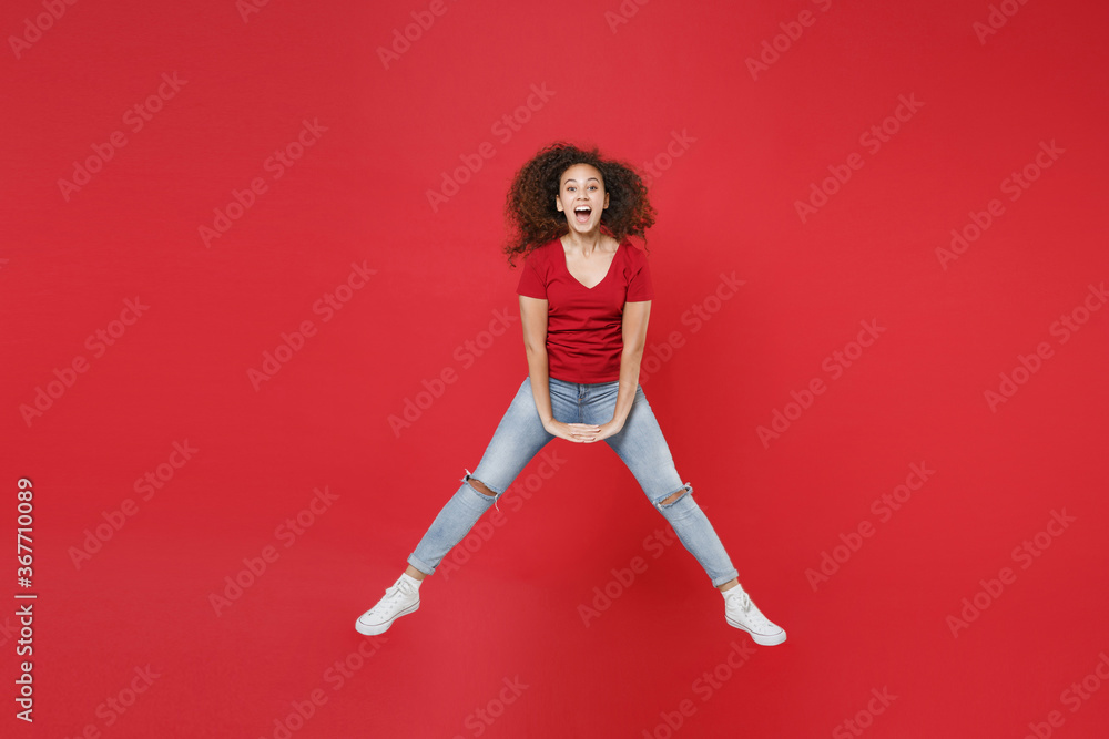 Full length portrait of excited young african american girl in casual t-shirt isolated on red wall background studio portrait. People lifestyle concept. Mock up copy space. Jumping spreading legs.