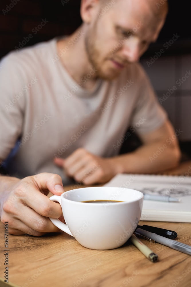 Selective focus of designer holding cup of coffee while working with sketchbook at table Selective focus of designer holding cup of coffee while working with sketchbook at table