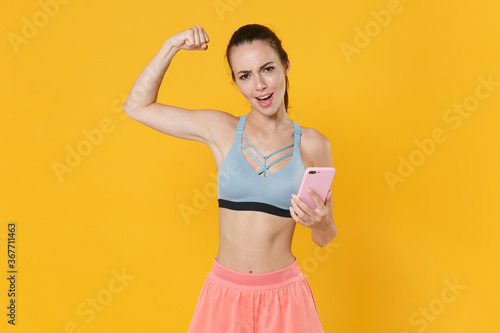 Strong young fitness sporty woman in sportswear working out isolated on yellow background. Workout sport motivation lifestyle concept. Mock up copy space. Using mobile phone, showing biceps, muscles. © ViDi Studio